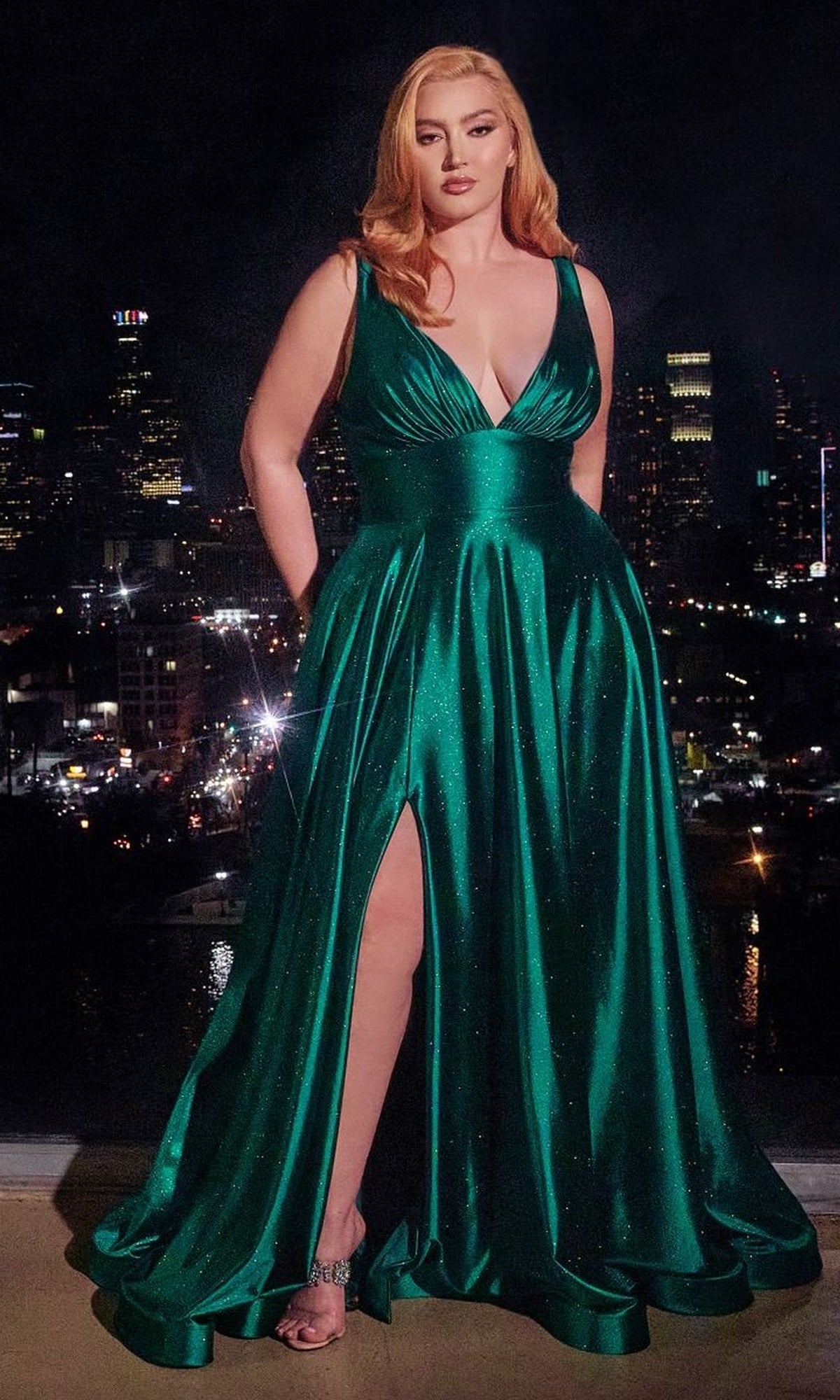 A woman wears the Plus Long Glitter Prom Dress: Ladivine CC2349C, a sleeveless emerald green plus-size gown with a high slit, standing indoors against a backdrop of a city skyline glowing at night.
